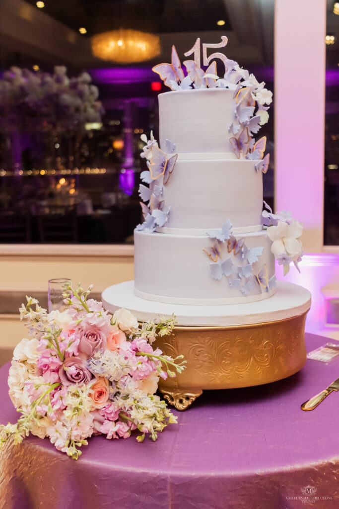 15 cake in lavender with lavender butterflies on top of a gold cake stand with a bouquet of pink and lavender roses on the left side.